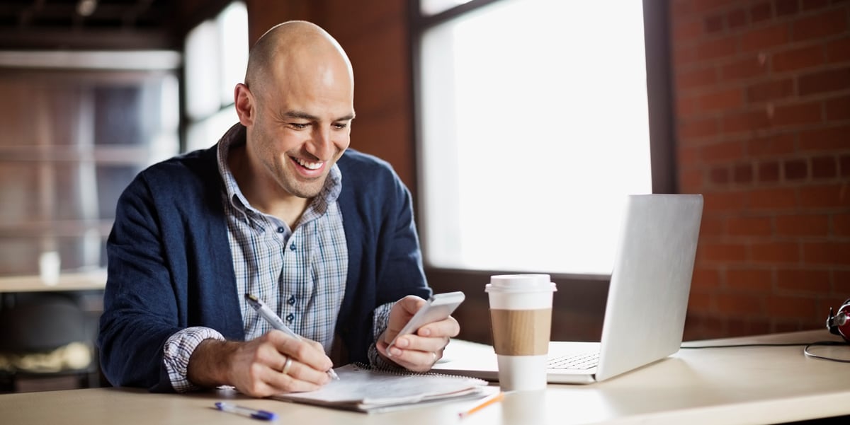 Smiling man writing at his desk with a coffee cup, laptop, in notebook in front of him